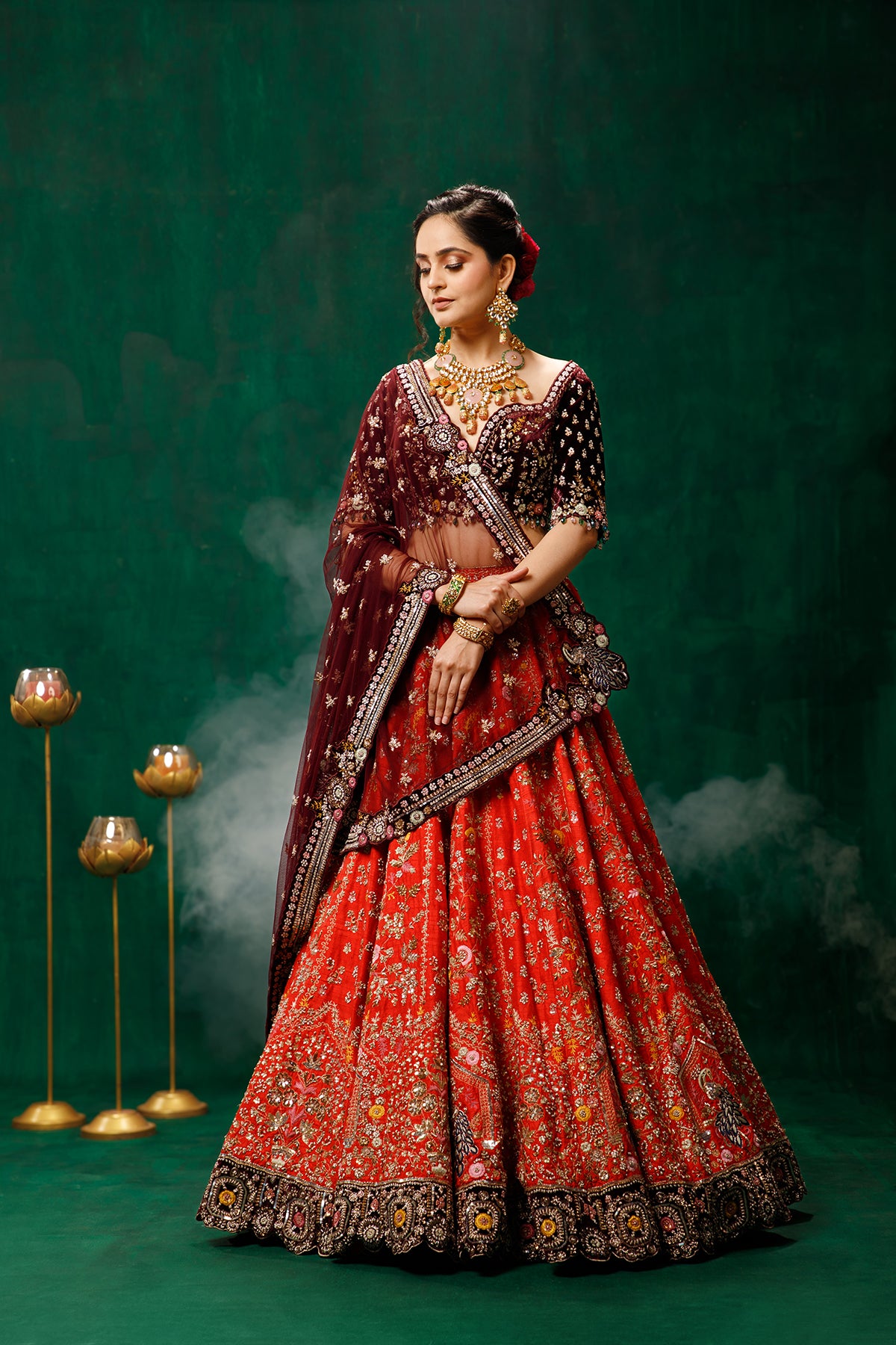 Woman in traditional red and maroon embroidered lehenga with jewelry against a dark green background