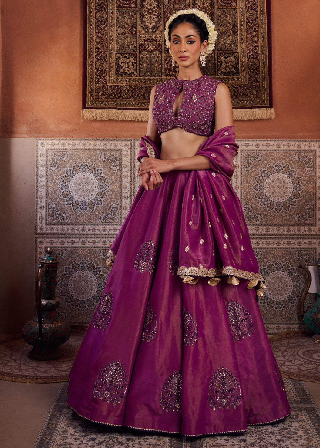 Woman in a traditional purple outfit standing in a decorated room with patterned walls and rugs.