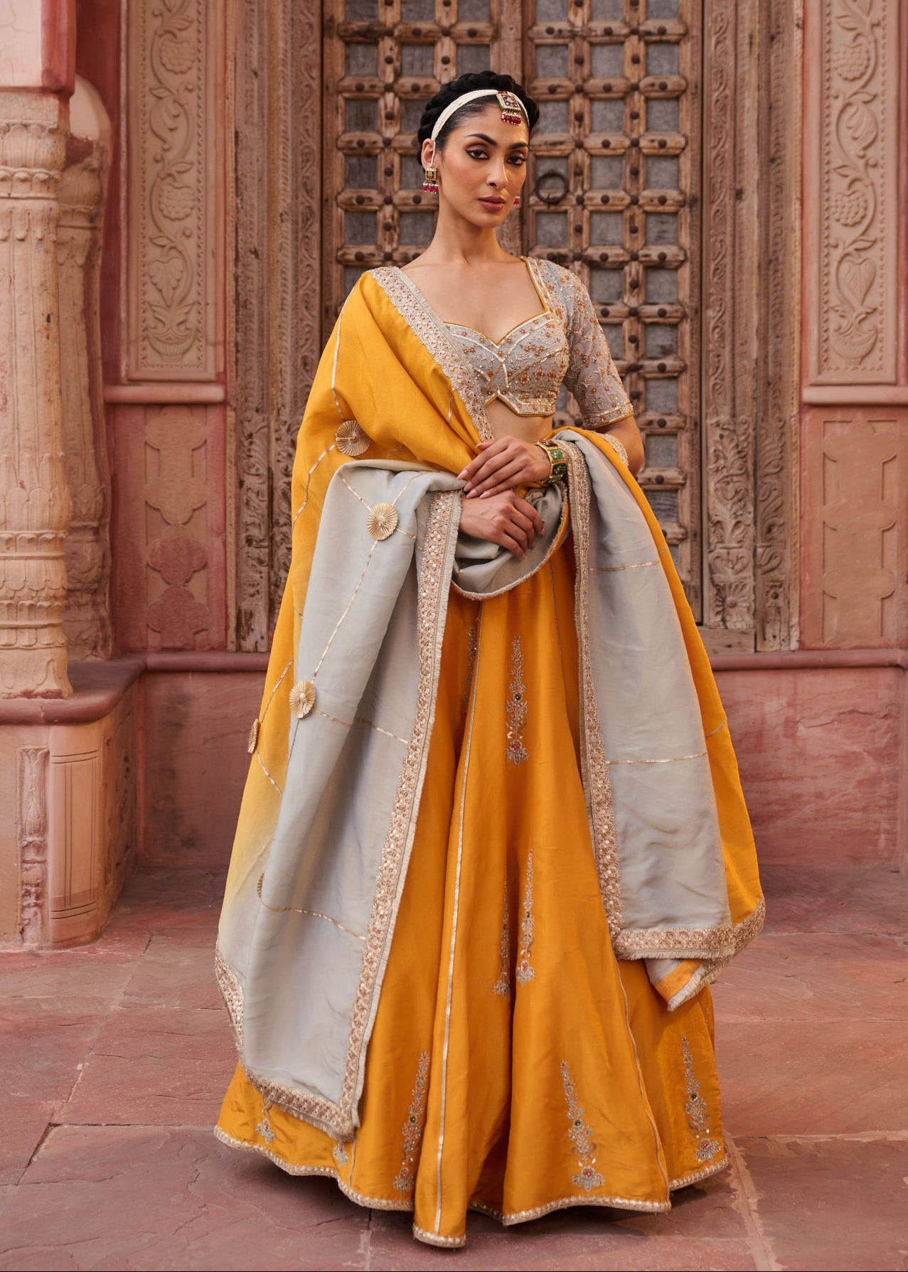 Woman in traditional yellow and gray outfit standing in front of an ornate wooden door.