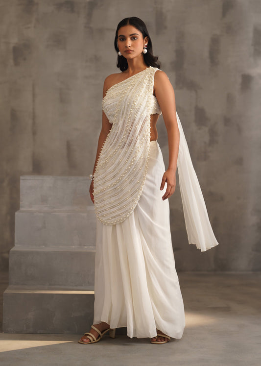 Woman in a white saree standing against a gray concrete wall.