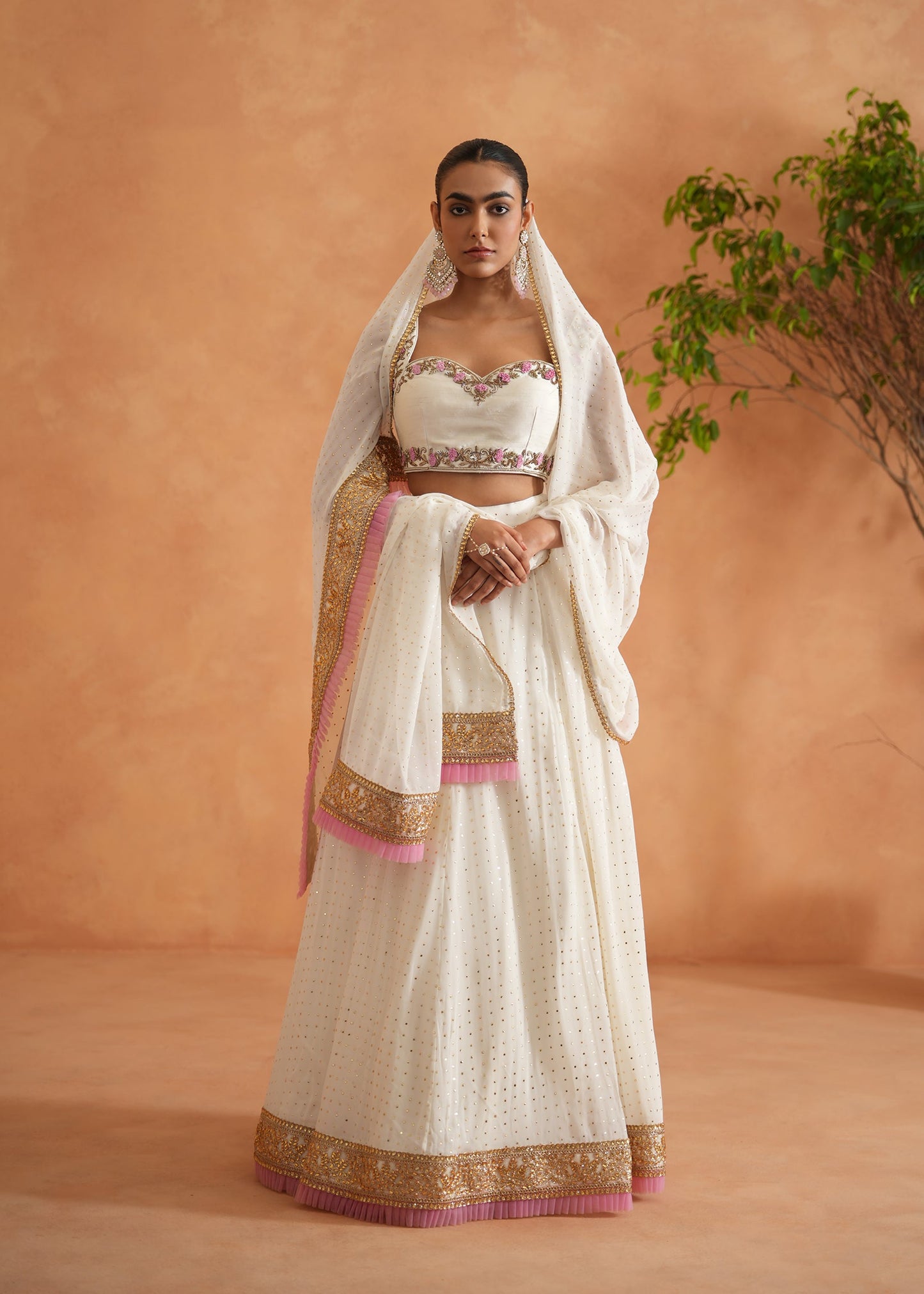 Woman wearing a traditional white saree with gold and pink details against an orange wall.