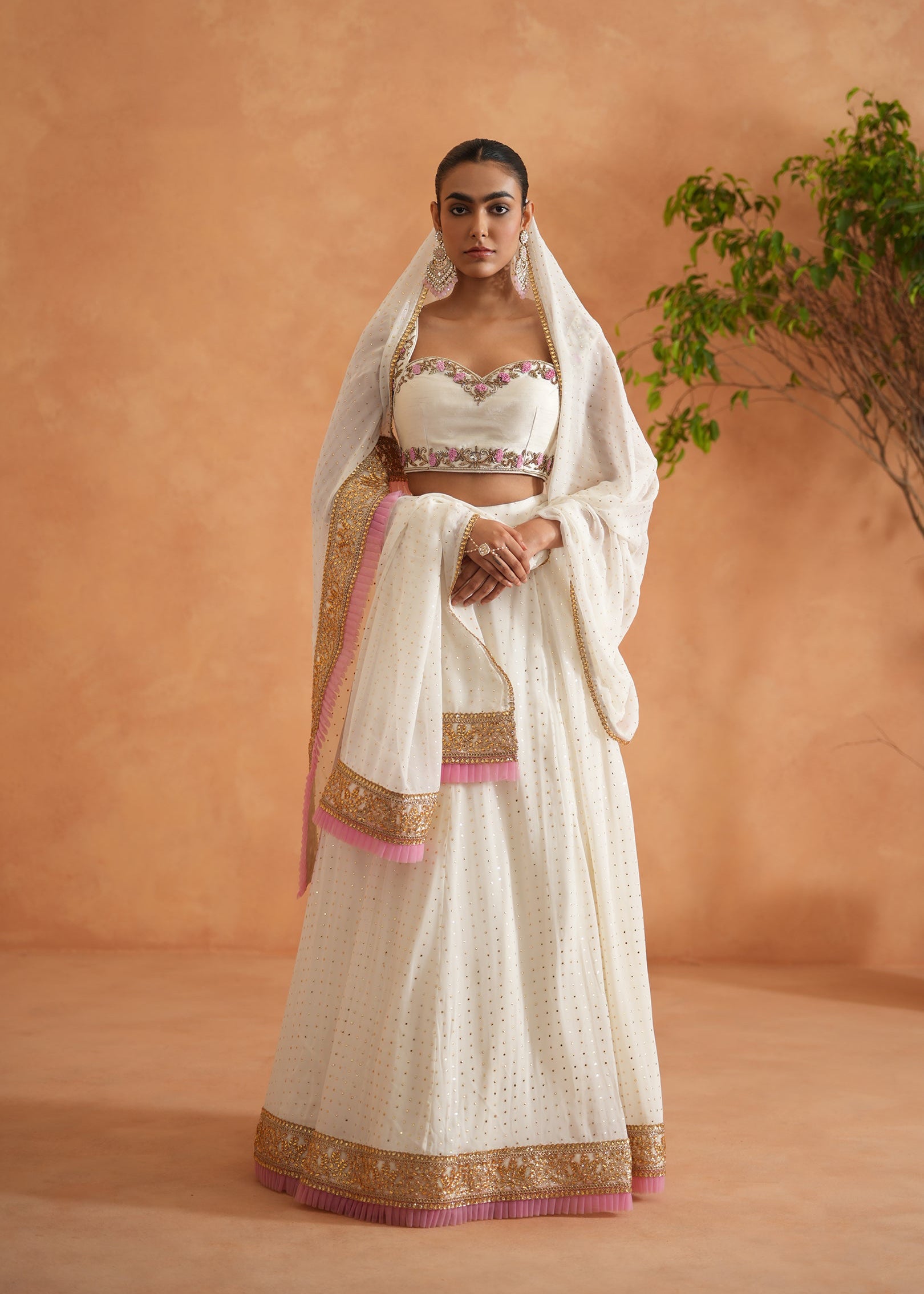 Woman wearing a traditional white saree with gold and pink details against an orange wall.