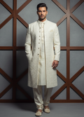 Man wearing a traditional cream sherwani against a geometric-patterned wall.