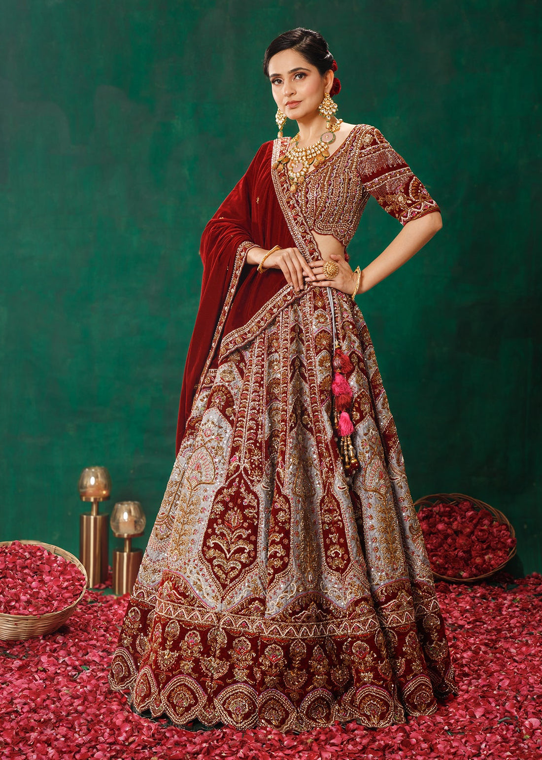 Woman in traditional maroon and gold embroidered outfit on a green background with red petals.