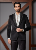 Man wearing a black suit with a white shirt and patterned tie against a wooden and gray background