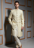 Man wearing a cream sherwani against a gray wall with wooden frame designs.