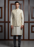 Man wearing a traditional cream kurta with an embroidered jacket against a gray wall.