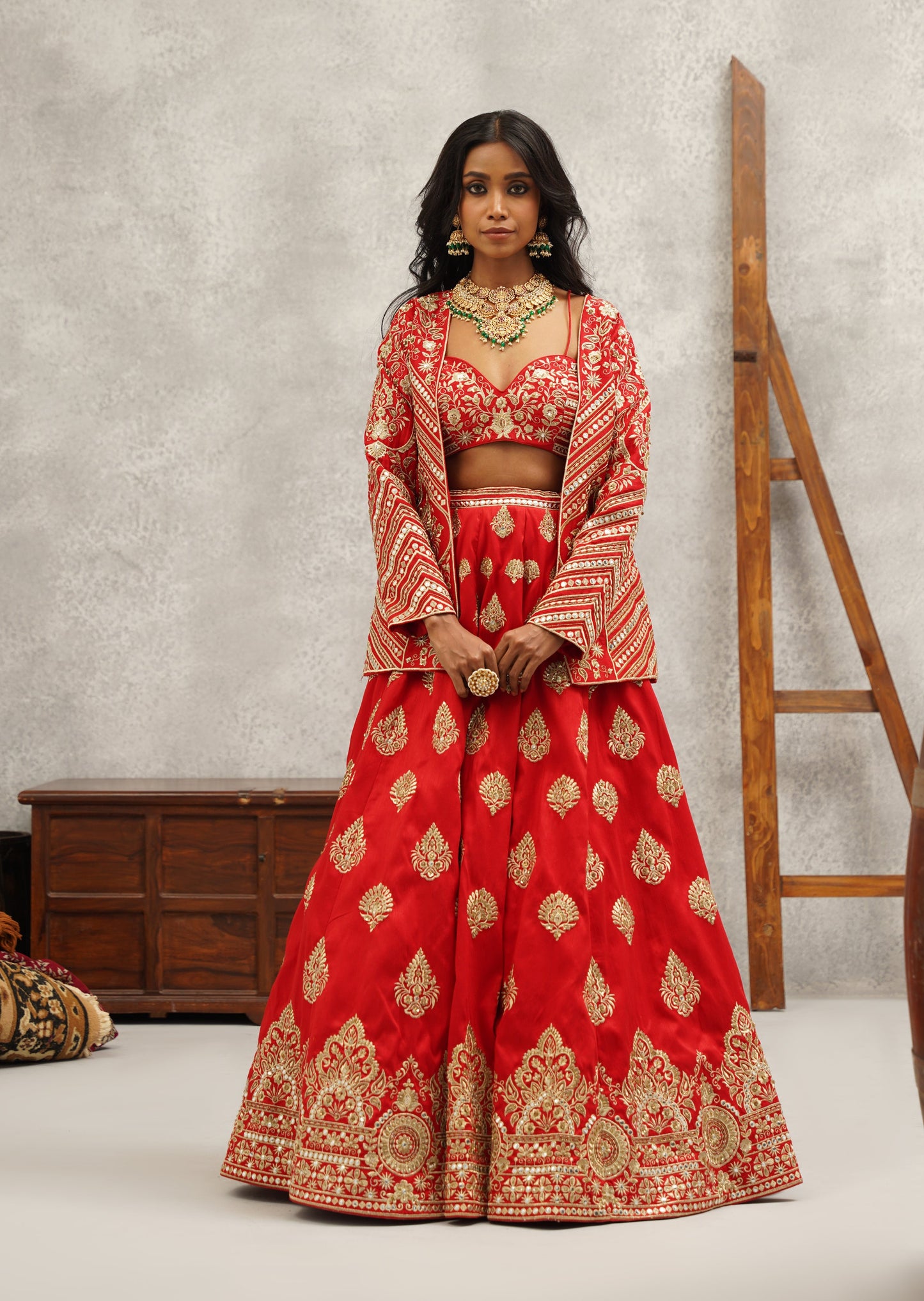 Woman in a red traditional outfit standing in a rustic setting with wooden furniture and a gray wall.