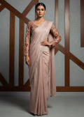 Woman wearing a light pink saree with floral blouse against a gray wall with wooden geometric patterns.