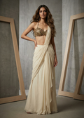 Woman wearing a gold top and white saree against a gray concrete wall.