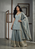 Woman wearing a light blue traditional outfit with gold embroidery in a minimalistic setting.