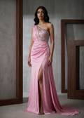 Woman wearing a pink one-shoulder evening gown with a high slit, standing against a neutral background.