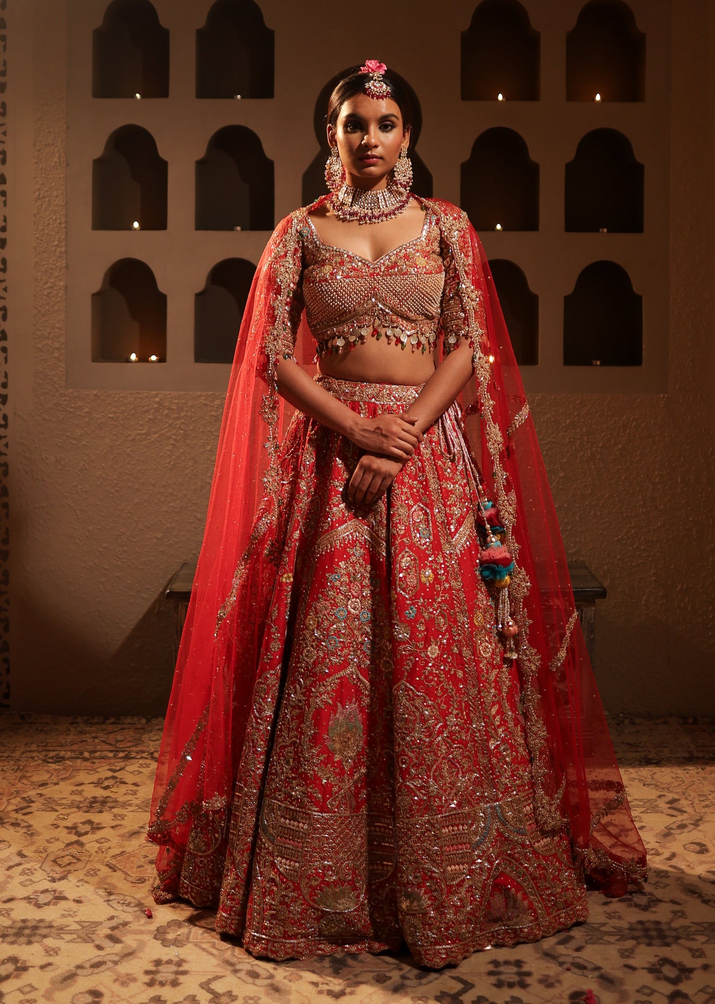 Woman in traditional red and gold embroidered outfit standing in a dimly lit room with arched windows.