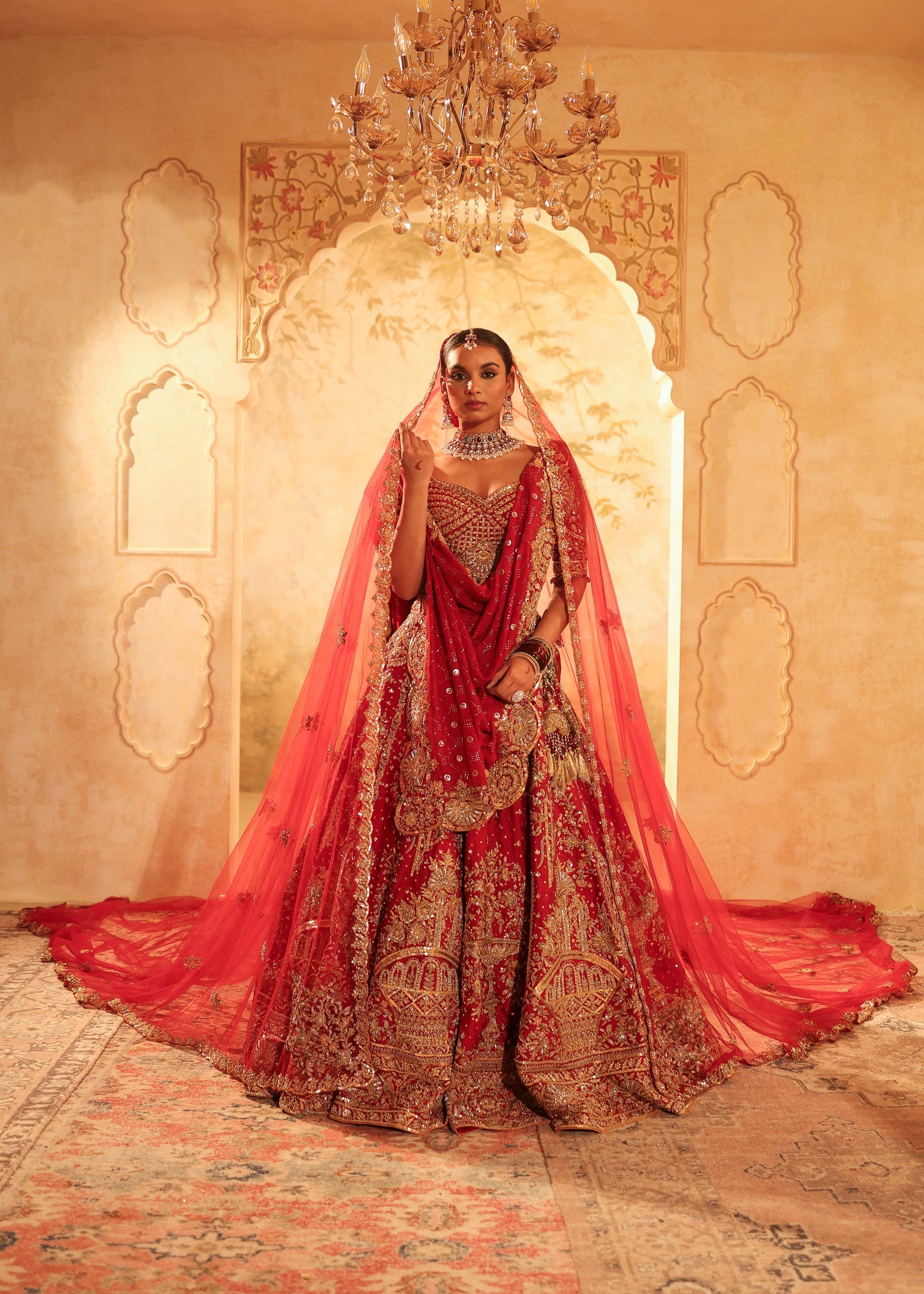 Woman in a red and gold traditional outfit standing in a decorated room with a chandelier.