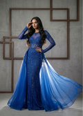 Woman in a blue evening gown with sheer overlay against a gray wall.