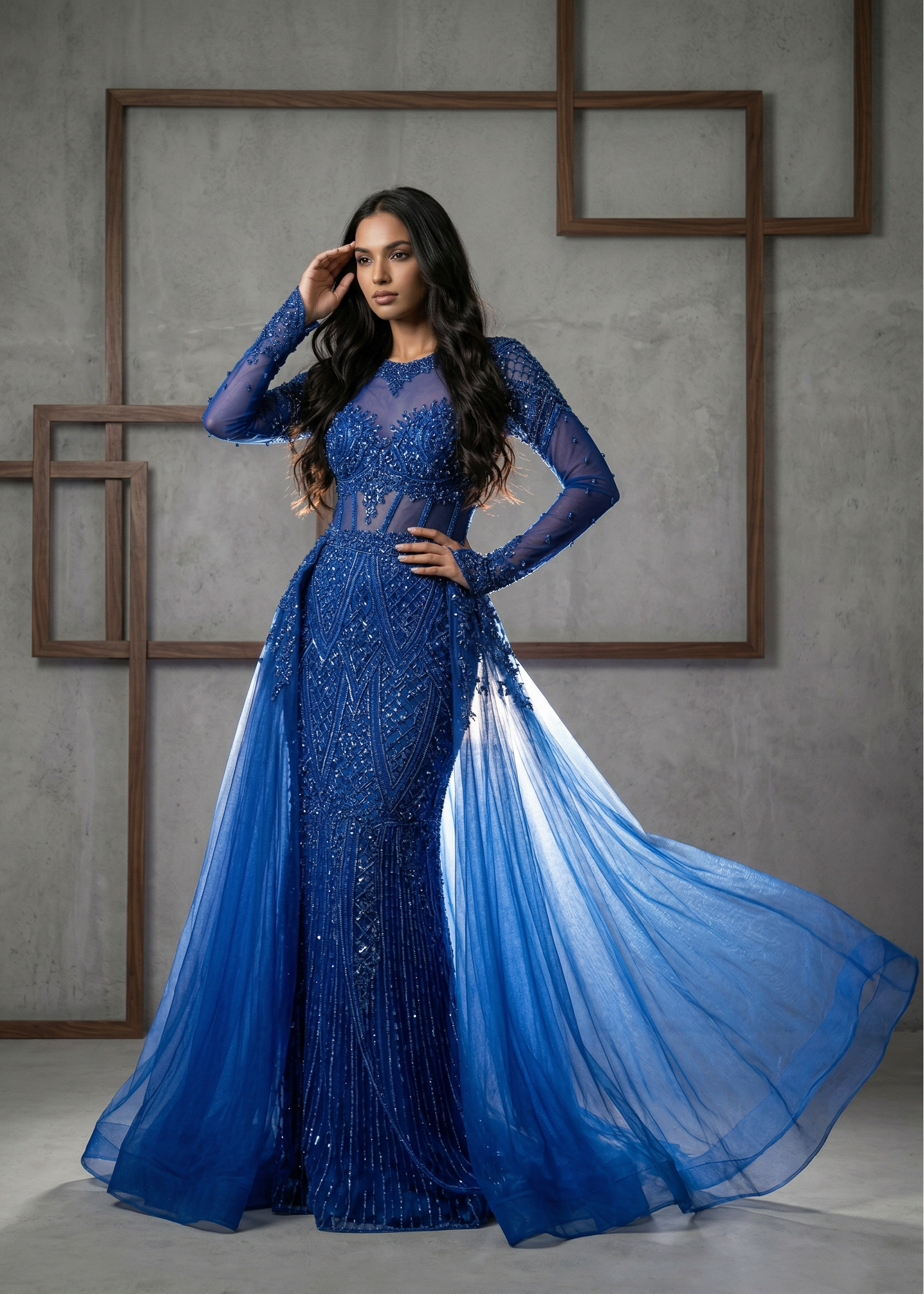Woman in a blue evening gown with sheer overlay against a gray wall.