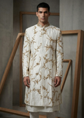 Man wearing a white embroidered sherwani against a gray wall with wooden frames.