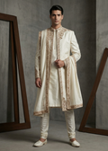 Man wearing a traditional cream sherwani with gold embroidery against a gray background