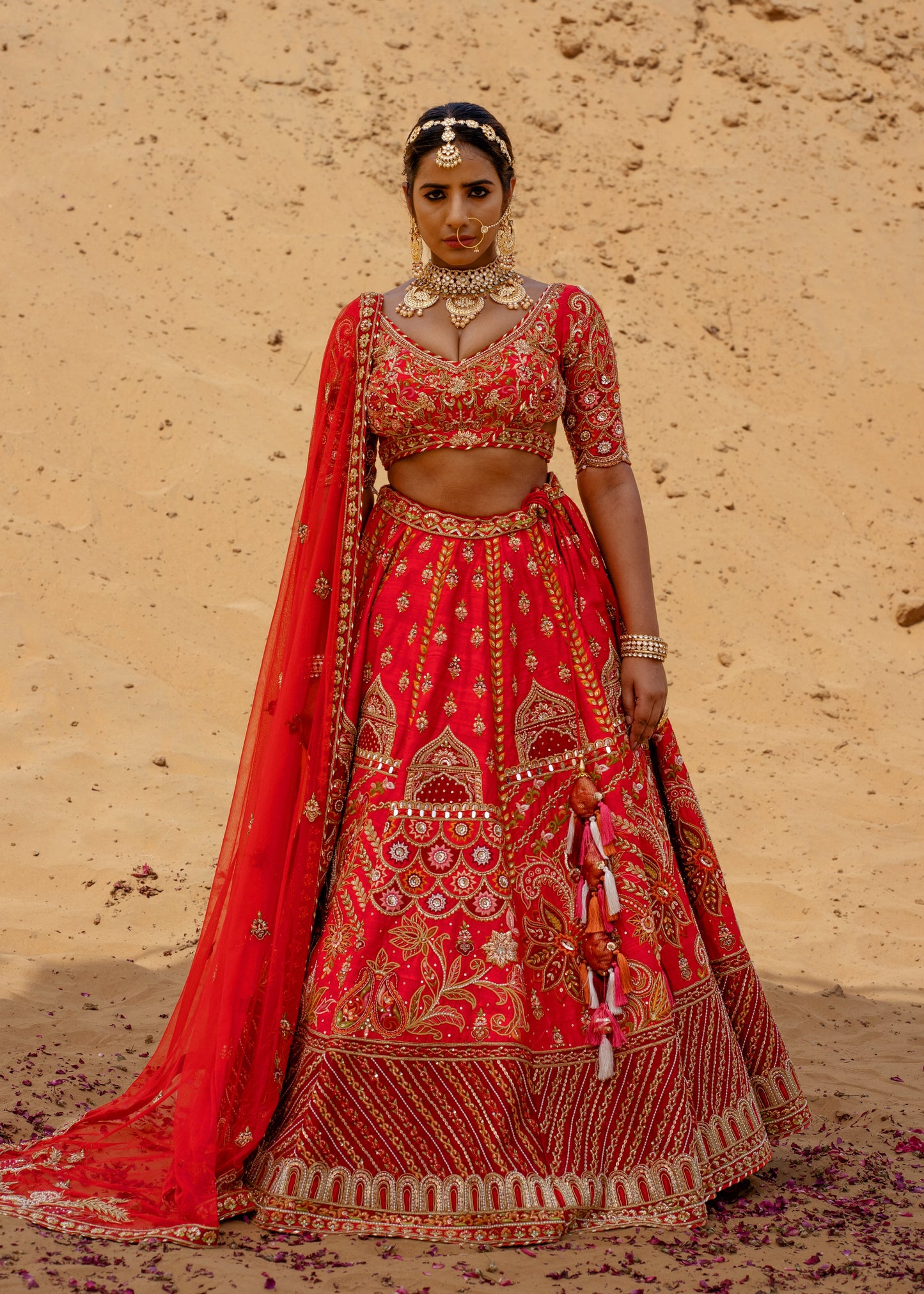 Woman in a traditional red embroidered outfit against a beige sandstone wall.
