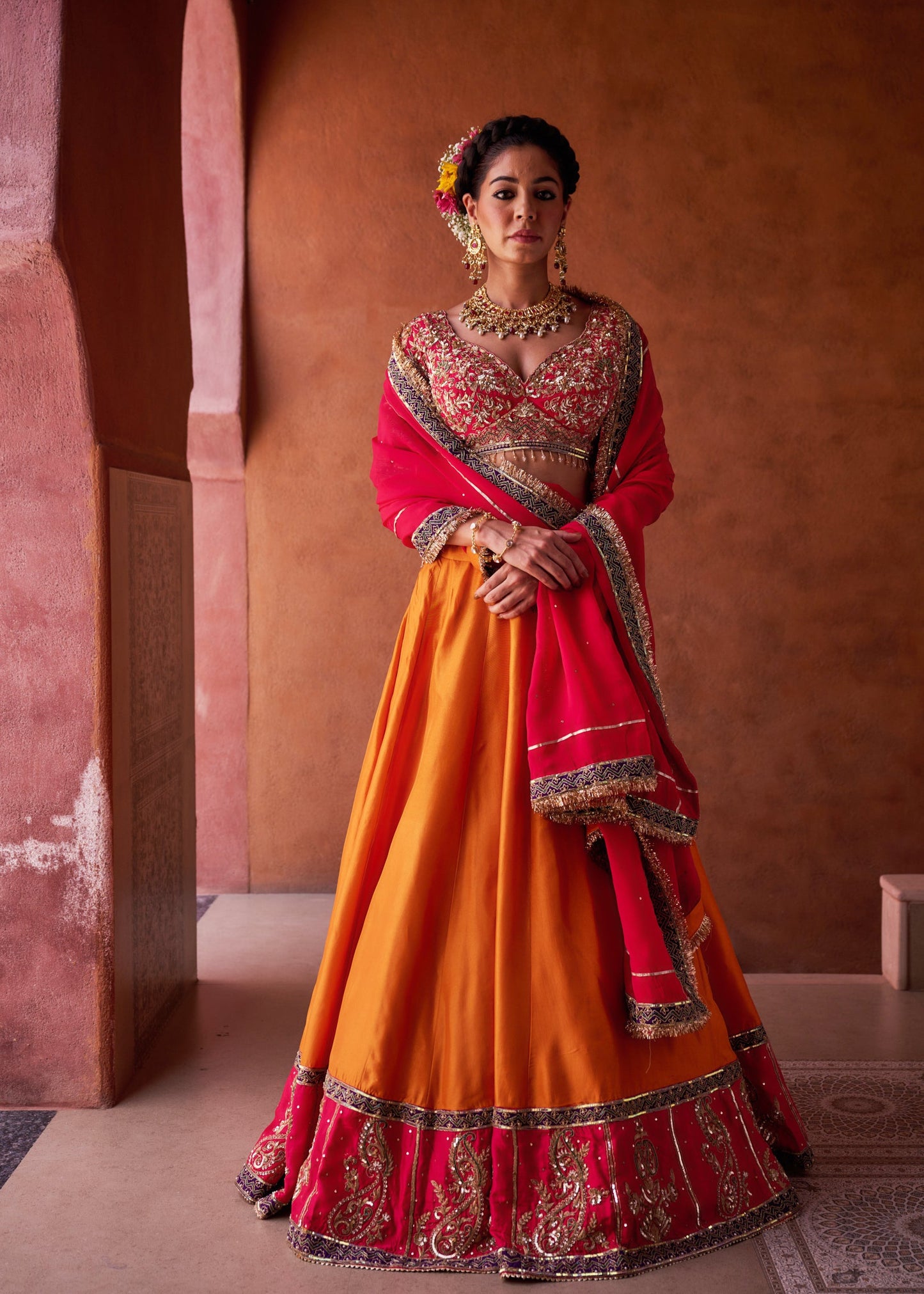 Woman in a traditional red and orange outfit with intricate patterns against a warm-toned wall.