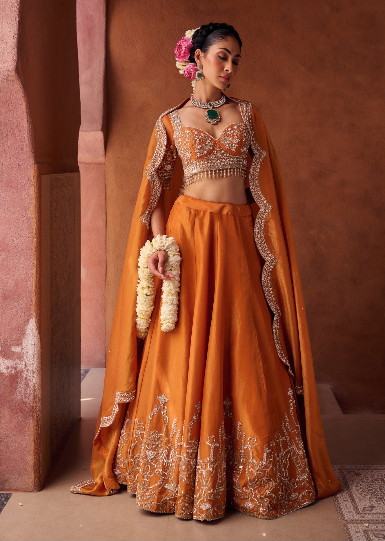 Woman in an orange traditional outfit with a cape against a warm-toned wall.