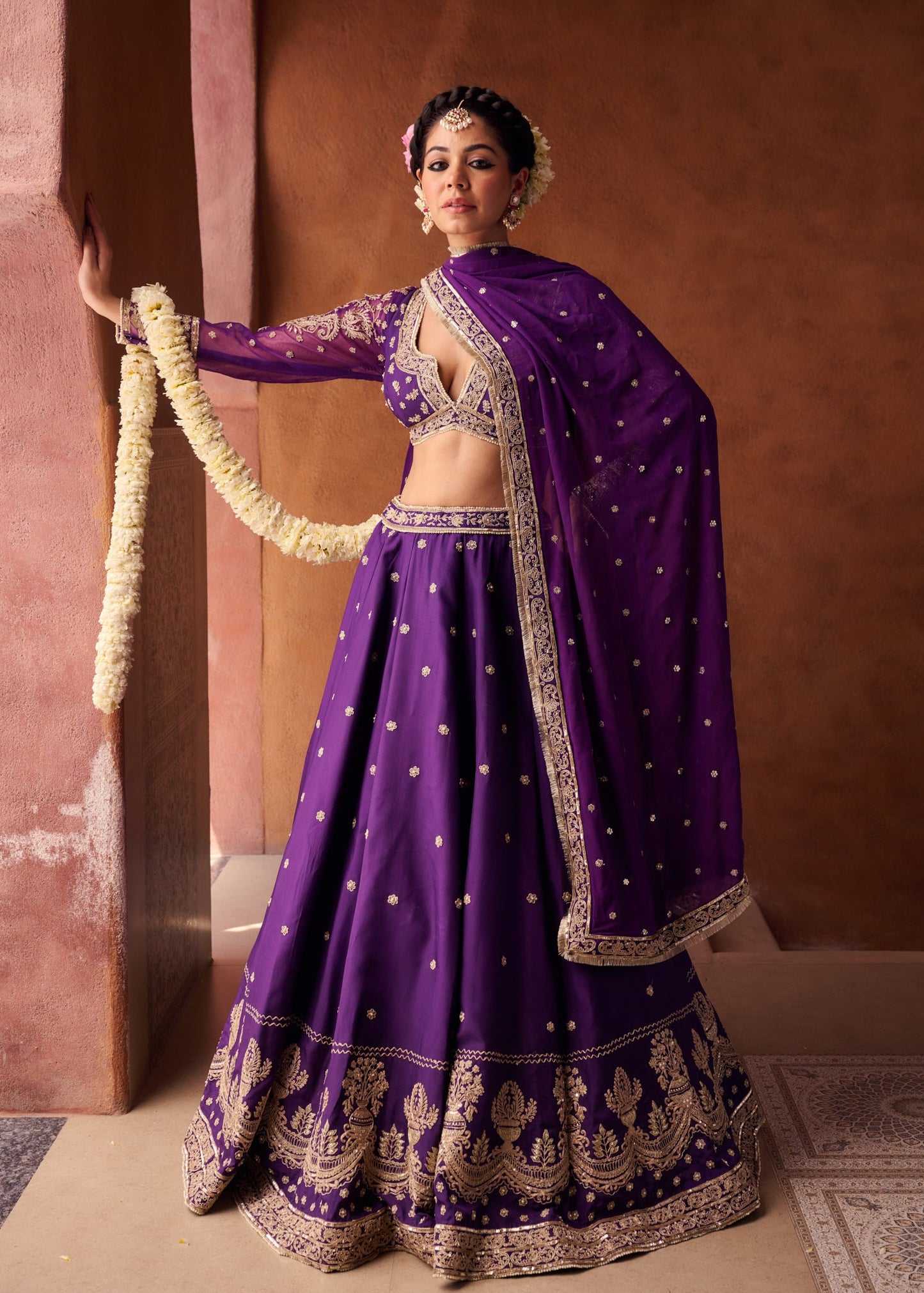 Woman in a traditional purple and gold outfit against a warm-toned wall.