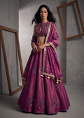 Woman wearing a traditional purple embroidered outfit with gold details against a gray background.