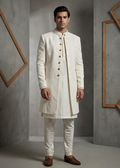Man wearing a traditional white sherwani against a gray wall with geometric wooden frames.